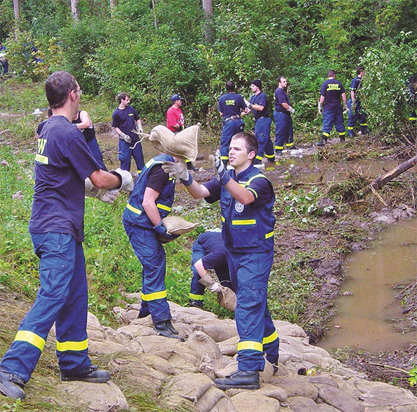 Das Foto zeigt Einsatzkräfte des Technischen Hilfswerks, die eine Kette gebildet haben und sich Sandsäcke zurwerfen. Im Hintergrund ein kleiner Bachlauf, der von den Einsatzkräften durch die aufgestapelten Sandsäcke blockiert wird.