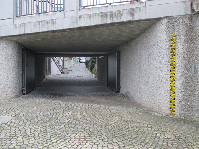 Auf der Abbildung ein eine massiv befestigte Fußgängerunterführung mit integriertem Hochwasserschutztor zu erkennen. Eine Pegelmesslatte zeigt im Ereignisfall den Wasserstand an. Ein gepflasterter Boden soll die Versickerung begünstigen.